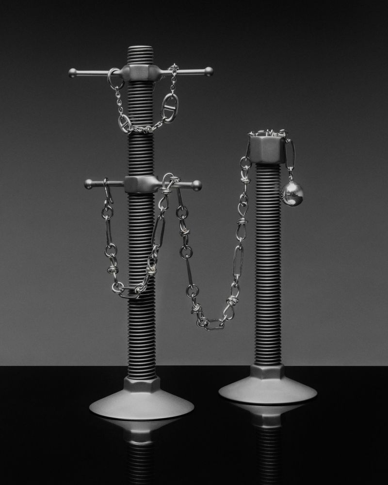 Two silver-tone metal threaded bars with chains draped across them against a grey background in a studio shot. Jewellery advertisement.