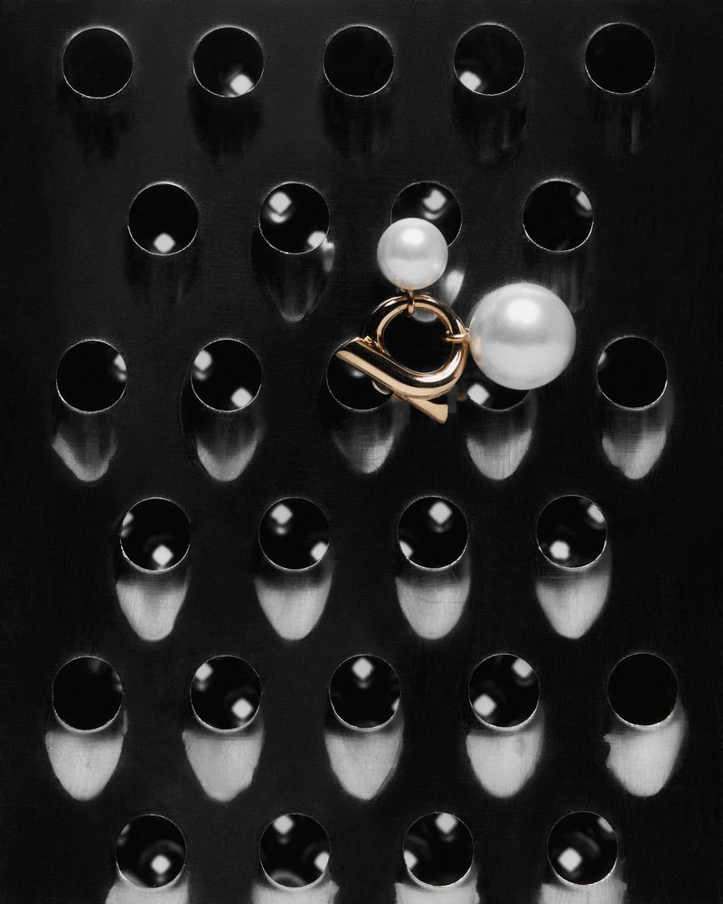 Overhead shot of gold and pearl earring on a black metal cheese grater, pearls reflecting light in a dark, minimalist studio product photo.
