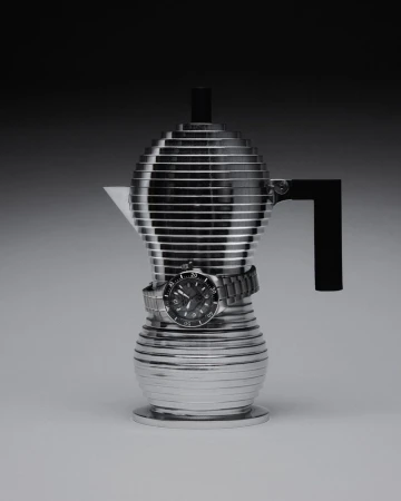 Sculptural tiered silver espresso pot wrapped with a luxury wristwatch against a dark gradient studio background.
