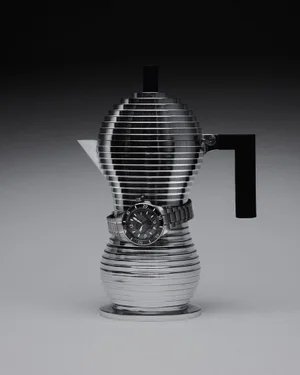 Sculptural tiered silver espresso pot wrapped with a luxury wristwatch against a dark gradient studio background.
