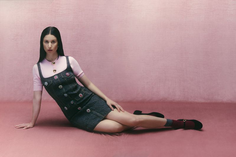 Woman in a denim dress posing against a pink backdrop. She's lying down, looking at the camera. The scene features a simple and clean aesthetic.