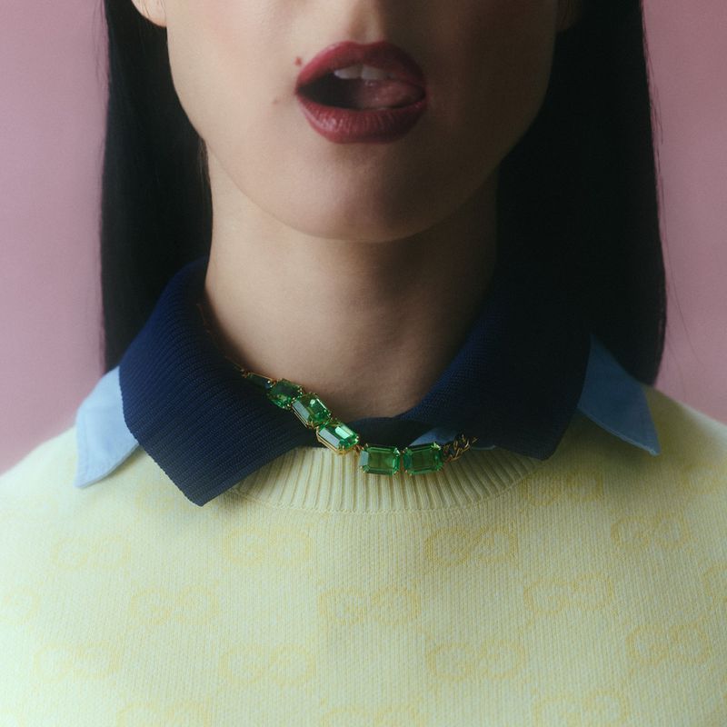 Close-up portrait of Francisca Valenzuela wearing an emerald gemstone necklace, navy collar, and pale yellow sweater on a pink backdrop.