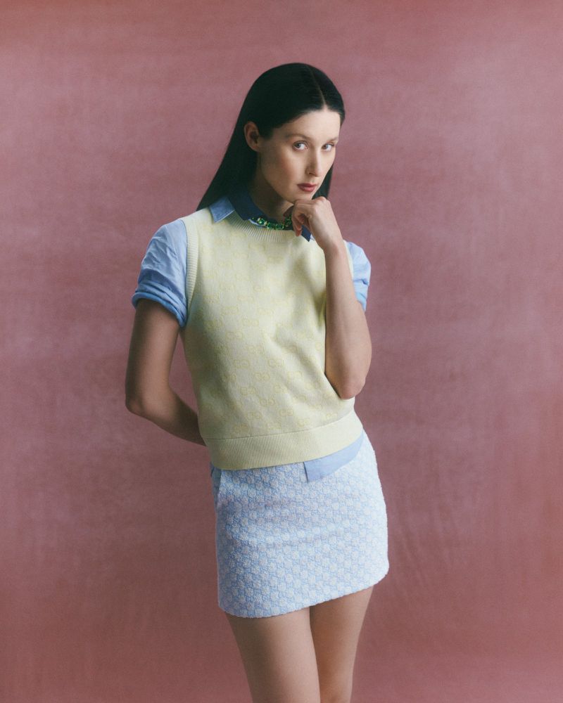 Young woman in preppy fashion poses against a textured pink background wearing a pale yellow sweater vest and blue and white mini skirt.