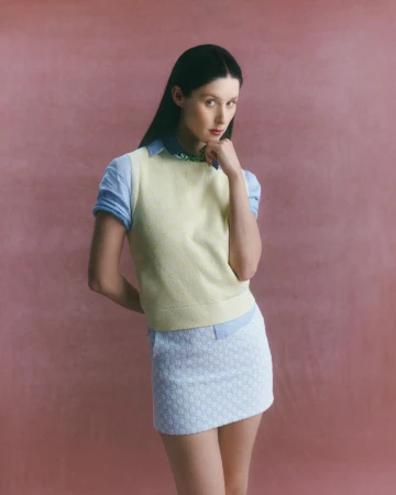 Young woman in preppy fashion poses against a textured pink background wearing a pale yellow sweater vest and blue and white mini skirt.