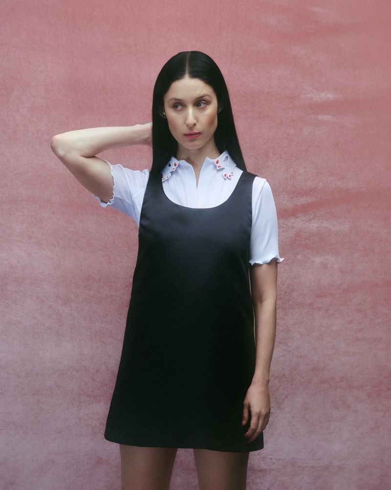 Brunette woman in an overall dress and white shirt posing against a pink textured backdrop in a fashion forward studio portrait.