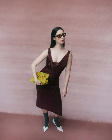 Francisca Valenzuela wearing a sleeveless burgundy cowl-neck dress, sunglasses, and white kitten heels against a pink textured backdrop.