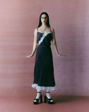 Francisca Valenzuela wearing a navy polka-dot slip dress with white lace and platform heels against a pink backdrop.