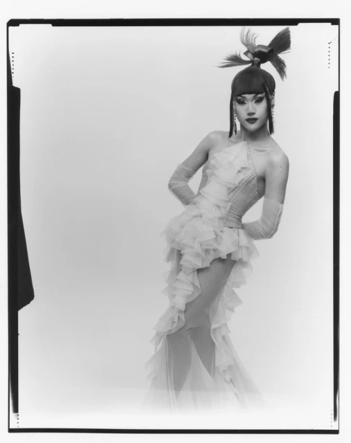 A person in an elaborate feathered headpiece and ruffled dress poses in a black-and-white studio shot with hands behind their back.
