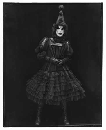 Monochrome portrait of an adult in a harlequin-inspired costume, featuring a ruffled dress, pointed hat, and distinct white and black makeup.