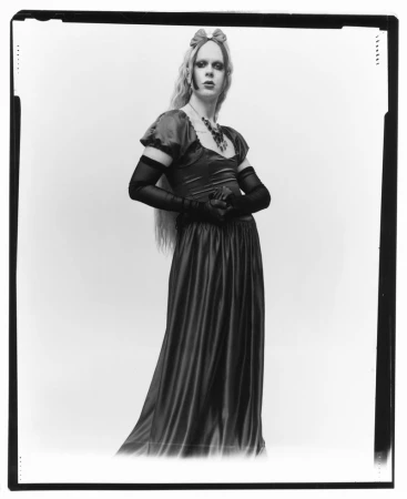 A full shot of a woman with long blonde hair in a dress with sheer sleeves and gloves, posing against a white backdrop.