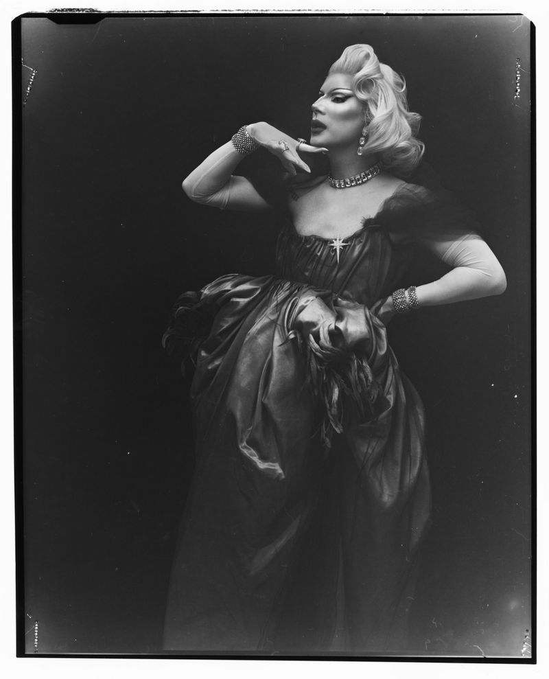 Drag queen in a dramatic pose wearing a gown, jewelry and glamorous makeup in a studio with a dark background. Full length shot.