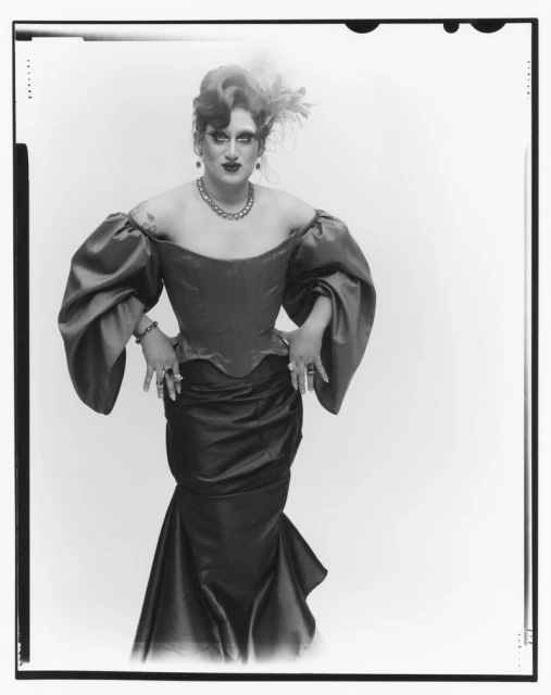 Drag queen poses in a black and white studio shot, wearing a red, off-the-shoulder dress with puffy sleeves, jewelry, and elaborate makeup.
