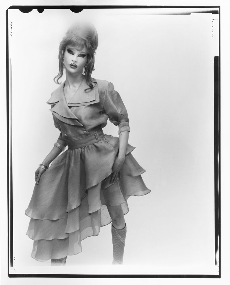 A person in a layered blue dress and knee-high socks poses against a white backdrop in a fashion editorial style portrait.