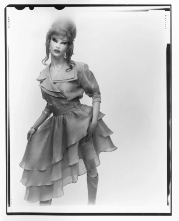 A person in a layered blue dress and knee-high socks poses against a white backdrop in a fashion editorial style portrait.