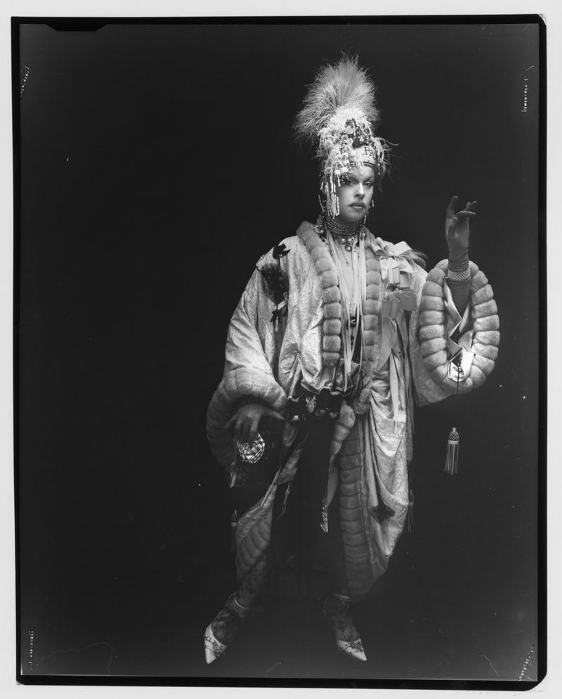 Full-shot of a performer in an ornate feathered headdress and costume with embellishments, posing in a studio with a dark backdrop.