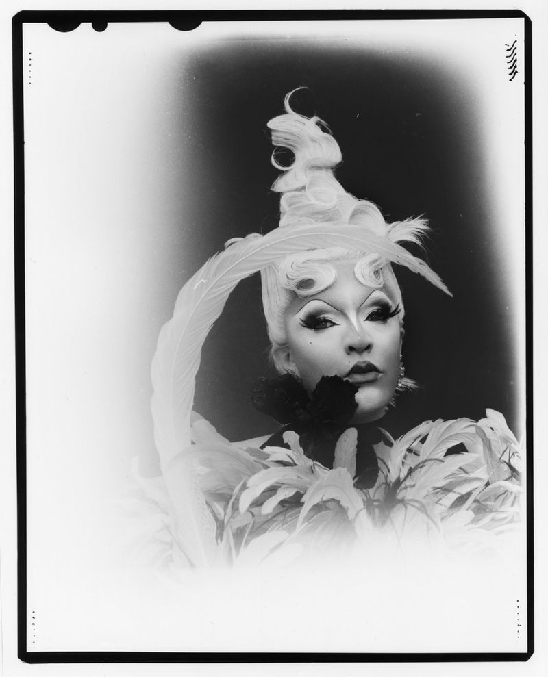 Dramatic black and white portrait of a person in elaborate feathered headwear, theatrical makeup, and a feathered collar.