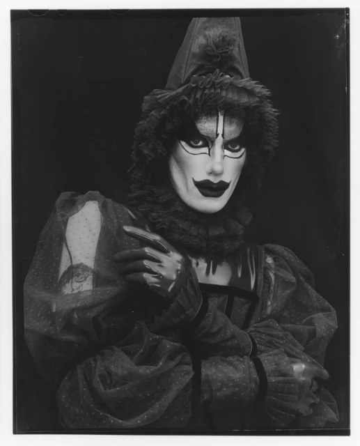 Dramatic black and white portrait of a person in theatrical makeup, wearing a polka dot outfit and pointed hat against a dark background.