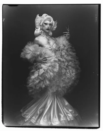 Elegant drag queen in a ruffled gown poses against a dark backdrop, showcasing glamour and artistry in a striking portrait.