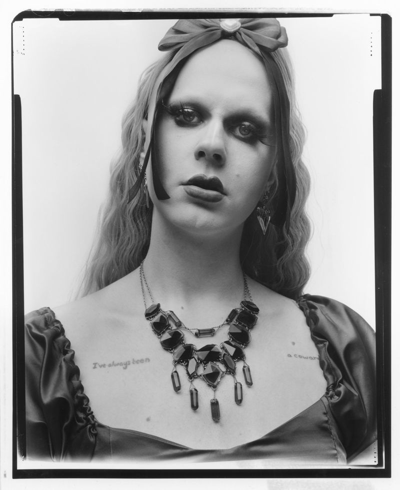 Dramatic portrait of a person with long hair, a red bow, and a statement necklace against a plain backdrop in a black and white photo.