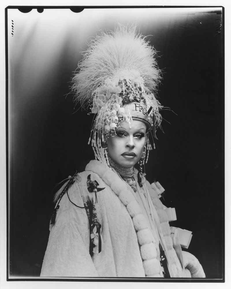 Striking black and white portrait of a person in an ornate feathered headpiece and fur coat against a dark backdrop.