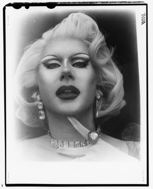Striking black-and-white portrait of a drag queen with platinum blonde hair, bold makeup, and extravagant jewelry, captured in a studio setting.