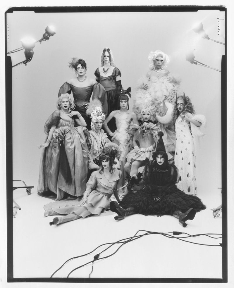 Group of eleven people in elaborate vintage-style costumes and theatrical makeup, posing in a bright studio setting with stage lighting.