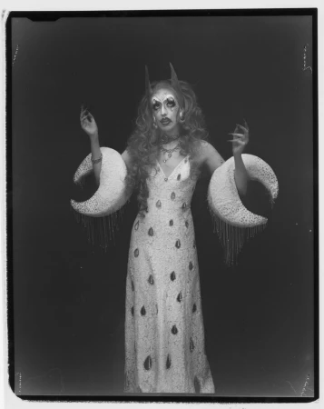 Drag performer in an embellished white gown with crescent moon arm pieces and dramatic makeup against a black backdrop.