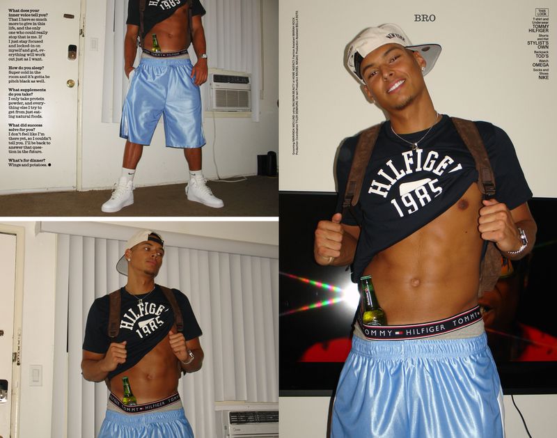 An adult male poses indoors, wearing a black Tommy Hilfiger t-shirt, light blue shorts, white sneakers and cap, and a brown backpack.