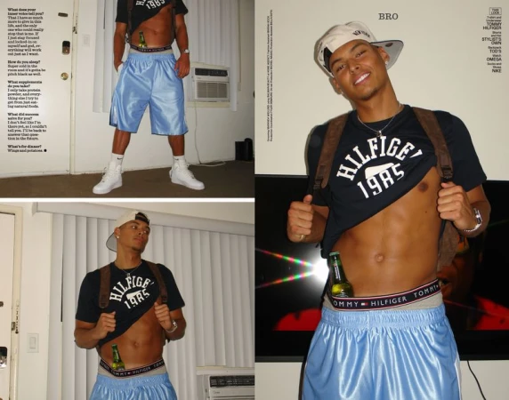 An adult male poses indoors, wearing a black Tommy Hilfiger t-shirt, light blue shorts, white sneakers and cap, and a brown backpack.