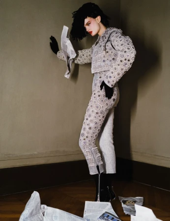 A young woman poses in a gray matador-style suit with black boots, gloves, and a feathered wig while holding and surrounded by newspaper.
