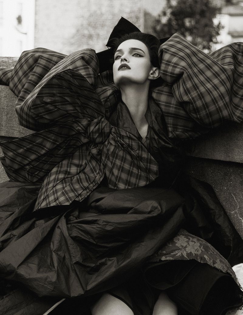Monochrome fashion shot: a model in a large plaid outfit, featuring puffed sleeves and a large bow, posing outdoors.