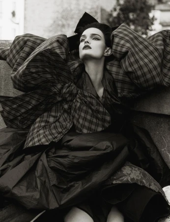 Monochrome fashion shot: a model in a large plaid outfit, featuring puffed sleeves and a large bow, posing outdoors.
