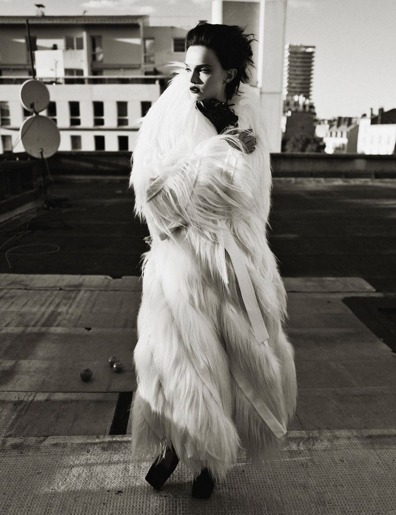 Monochrome portrait of a woman in a fur coat on a rooftop, with city buildings in the background, evoking high fashion and urban elegance.