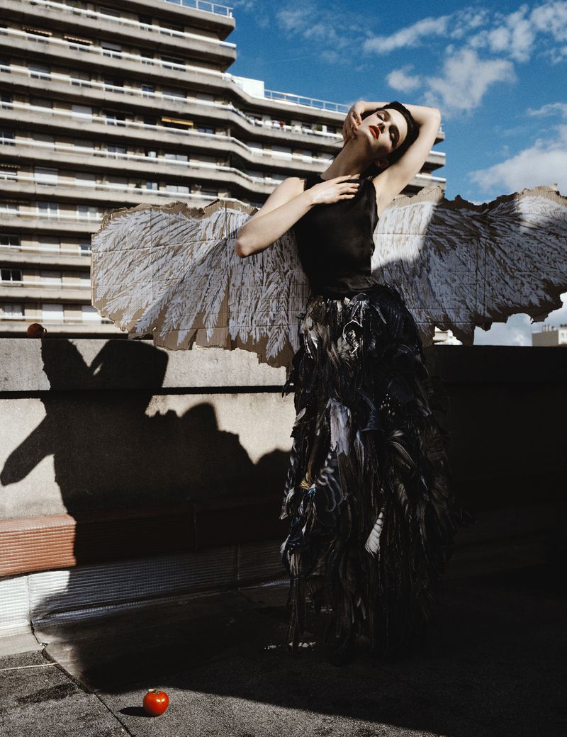Fashion shot of a slender woman with outstretched arms, wearing a black dress and large paper wings, posing against a building backdrop.