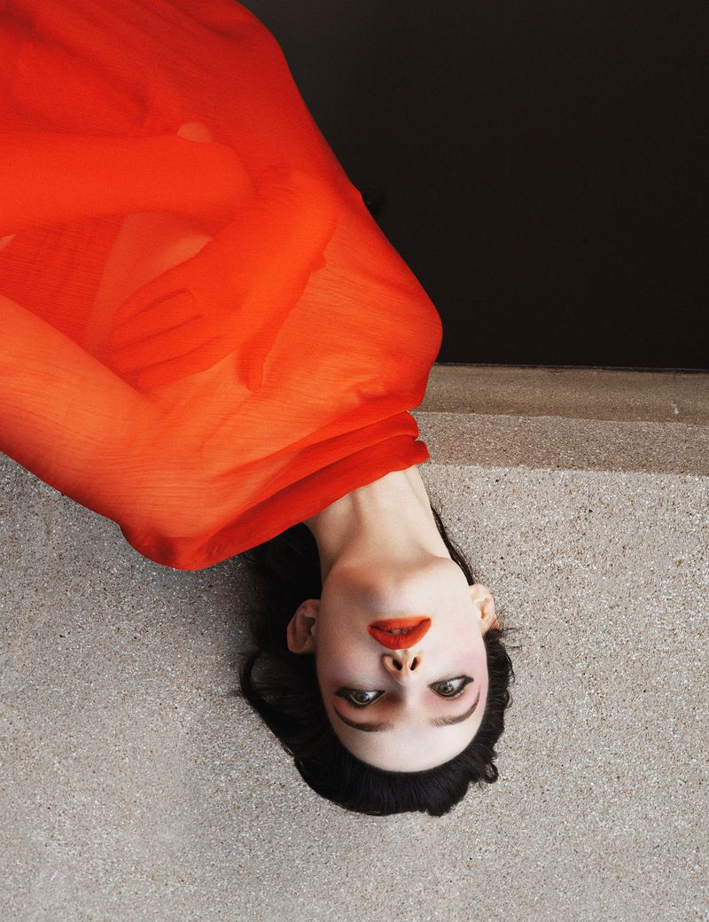 Portrait of a woman with dark hair wearing bright red lipstick and an orange translucent fabric, against a grey and black background.