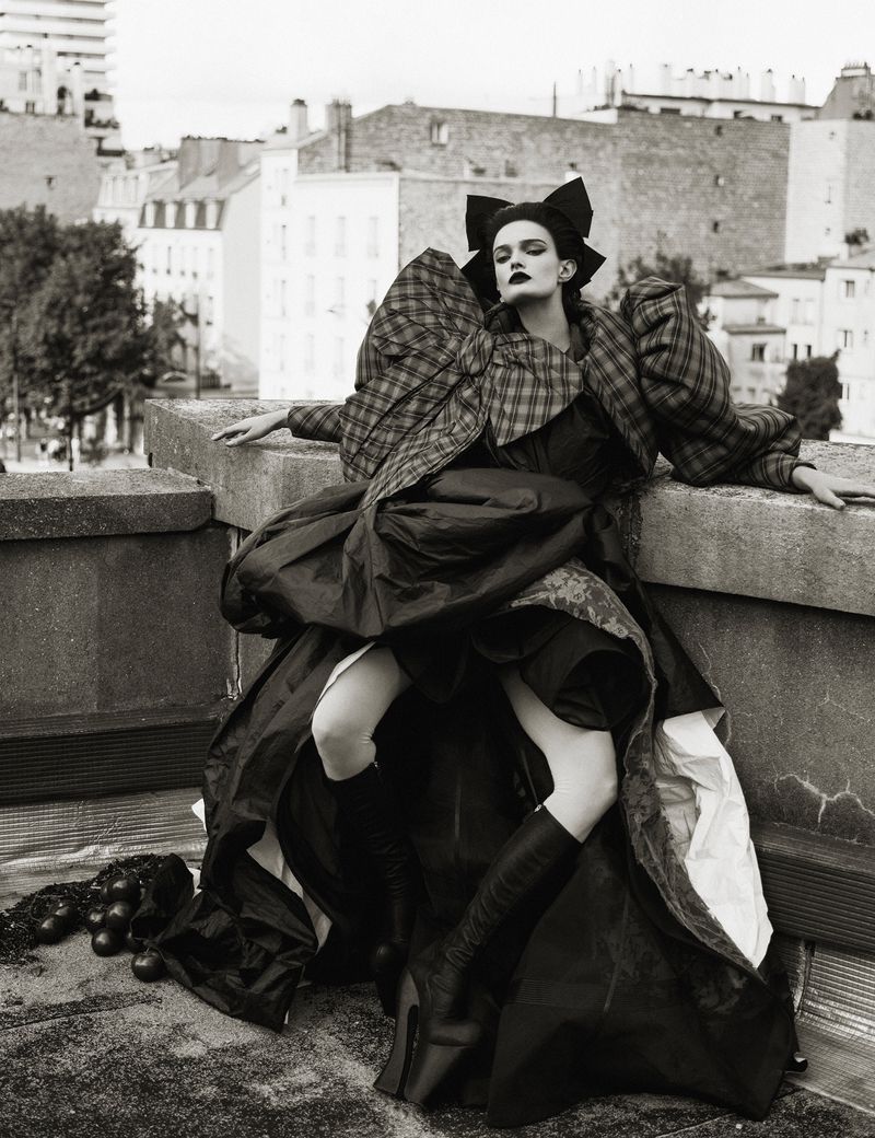 Fashion model in dramatic, oversized black dress and plaid jacket poses on rooftop with city buildings in the background.