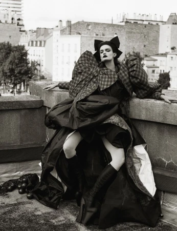 Fashion model in dramatic, oversized black dress and plaid jacket poses on rooftop with city buildings in the background.