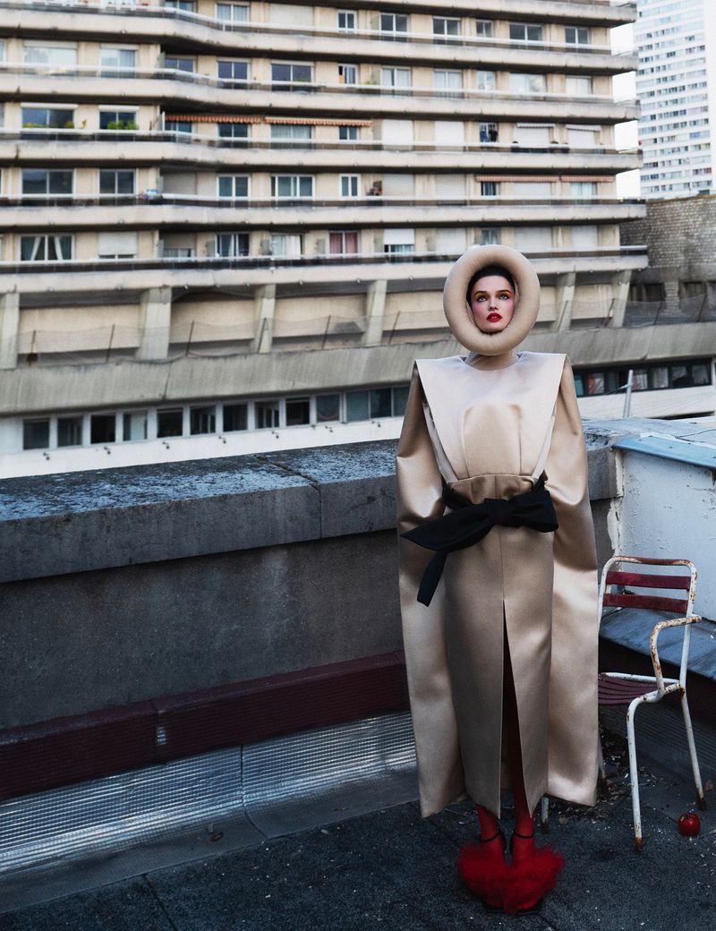 Woman in futuristic beige dress with black belt and red shoes stands on rooftop in front of brutalist buildings for fashion editorial.