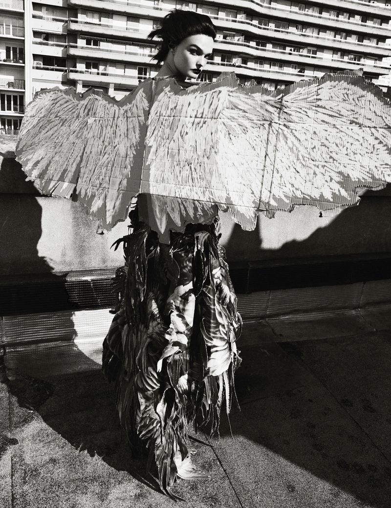 Monochrome outdoor fashion portrait of a model wearing feathered wings and garment, standing in front of a high-rise building in daylight.