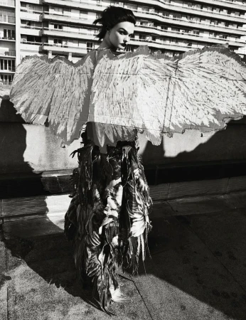 Monochrome outdoor fashion portrait of a model wearing feathered wings and garment, standing in front of a high-rise building in daylight.