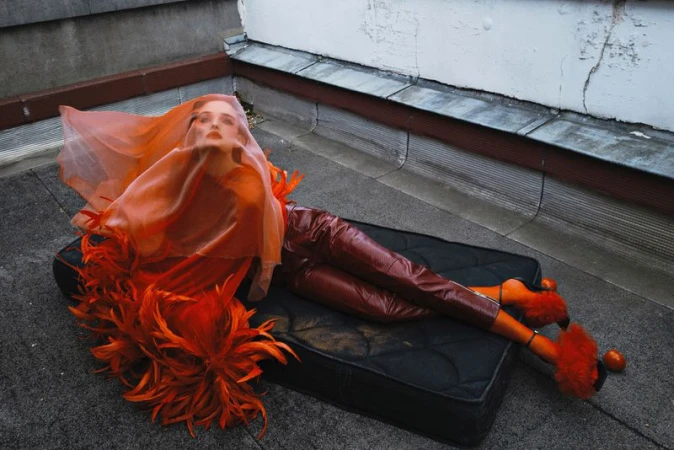 Woman in orange couture fashion with feathered accents and matching heels, laying on a dark surface in an urban environment.