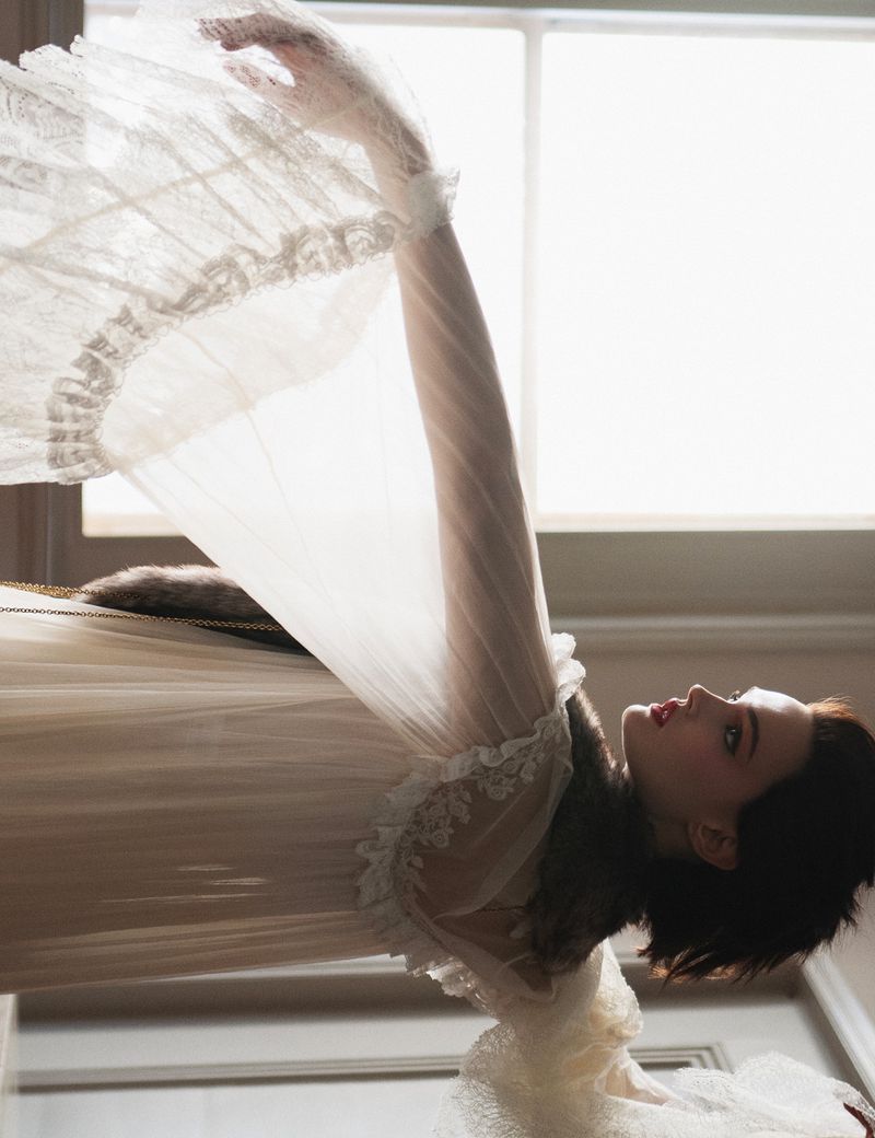Woman in sheer white gown poses with arms raised near window. Editorial fashion portrait of a female with brunette hair in indoor setting.