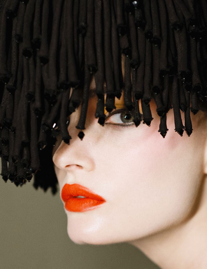 Close-up portrait of a fair-skinned adult woman with orange lipstick and a decorative black headpiece with beaded fringe.