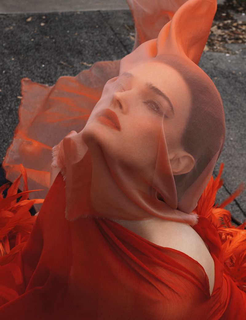 Woman with orange fabric and feathers poses outdoors, emphasizing face and fashion elements against a neutral background, shot with soft lighting.