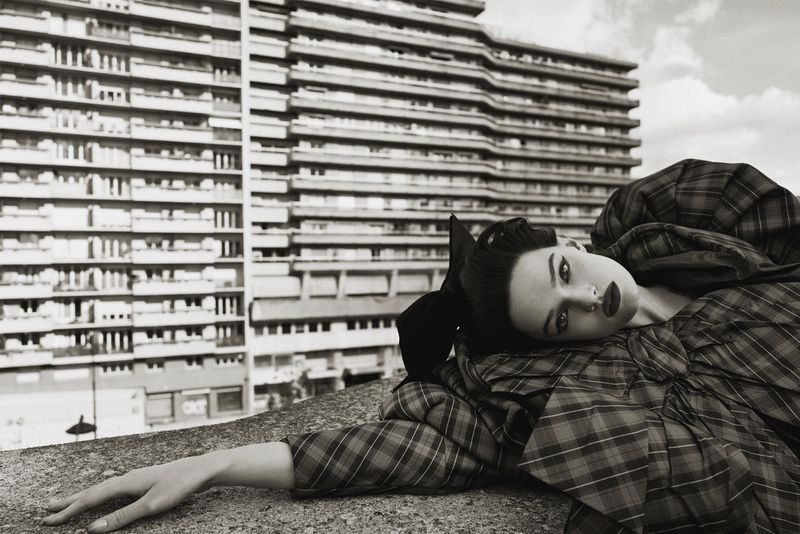 Fashion shot of a woman in a plaid dress, lying down with a modern, high-rise building in the background. Editorial fashion photography.