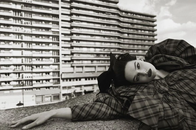 Fashion shot of a woman in a plaid dress, lying down with a modern, high-rise building in the background. Editorial fashion photography.