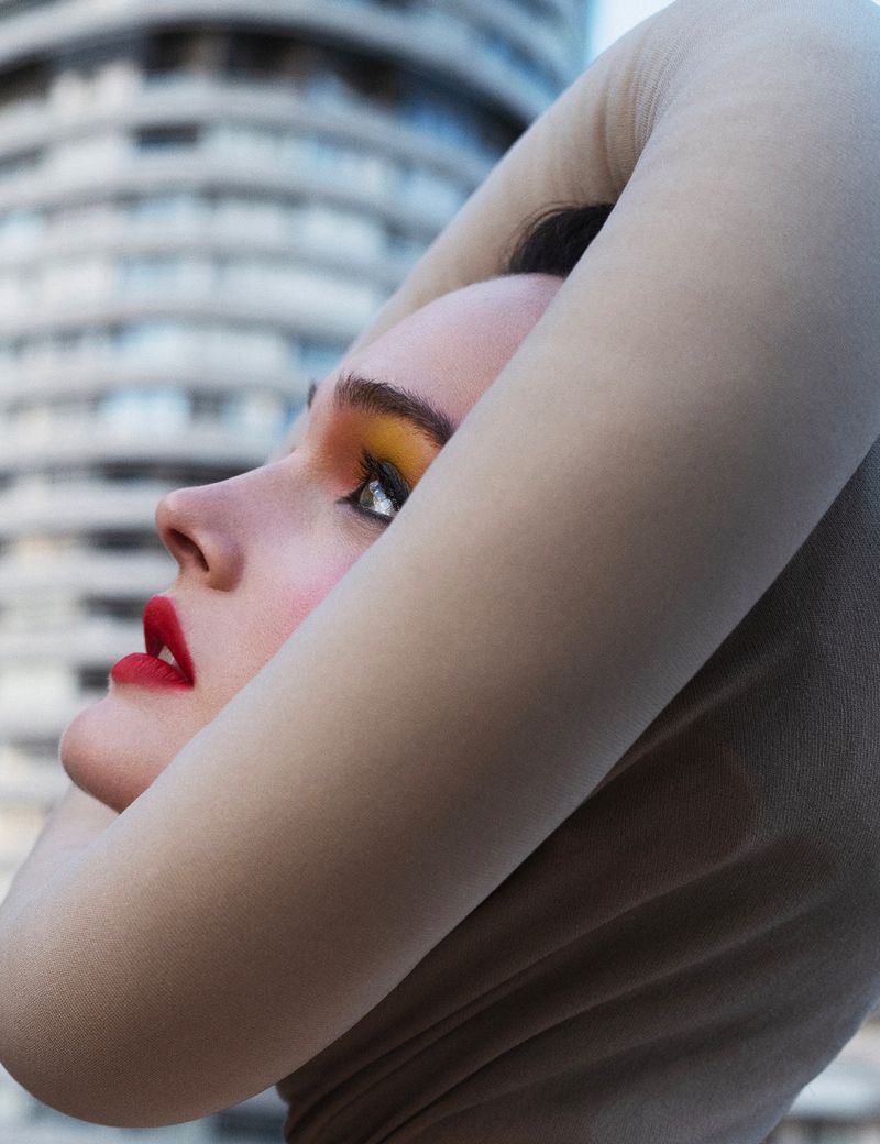 Woman in close-up profile with red lipstick and yellow eye makeup, wearing a beige top against a tall, modern urban building.