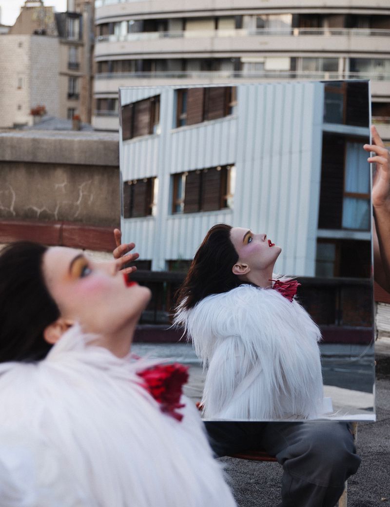 Brunette woman in a white jacket poses with a mirror on a rooftop with buildings in the background reflecting in the mirror.