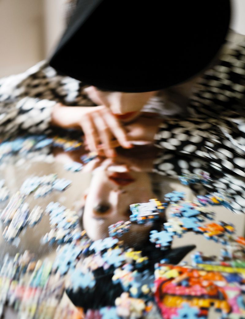 Abstract image of a woman reflected in a mirror with puzzle pieces scattered, wearing a black hat and spotted shirt.