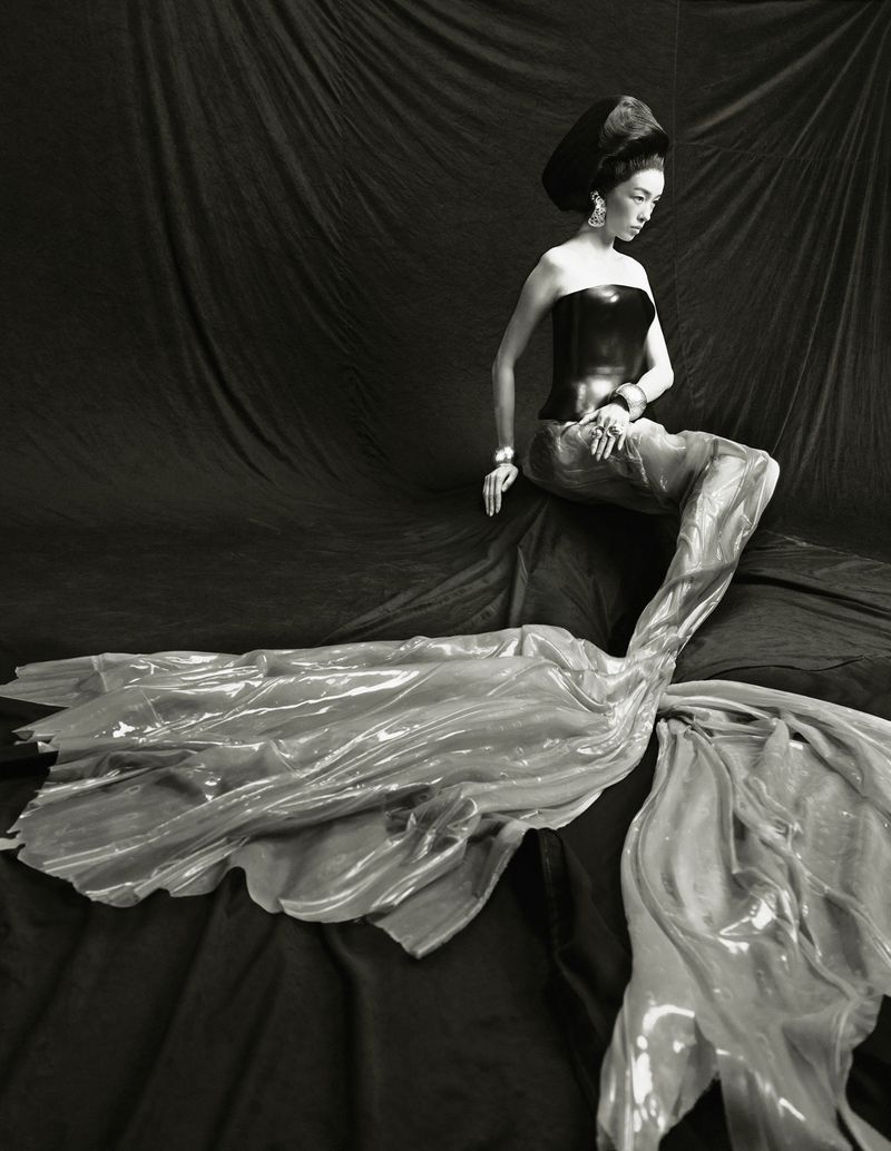 A fashionable woman with elaborate headwear and long, flowing skirt sits against a dark fabric backdrop in a black and white photograph.